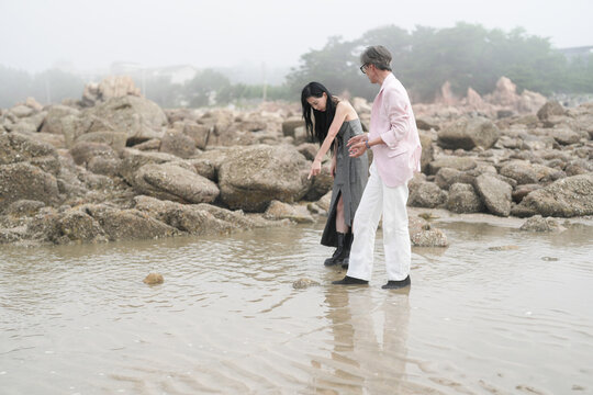In June 2025, a Korean man and woman in their late fifties are playing together along the rocky shoreline, stepping over jagged stones and enjoying the coastal scenery in Incheon Metropolitan City.