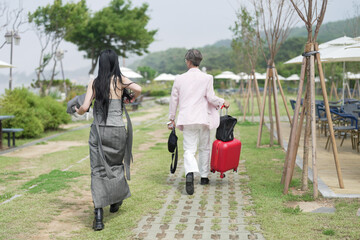 In June 2025, a Korean man in his late 50s walks ahead with a suitcase, followed by a woman of the...