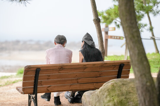 In June 2025, a Korean man and woman in their late fifties sit on a bench by the sea, chatting and smiling as they enjoy the breeze, ocean view, and calm surroundings in Incheon Metropolitan City.