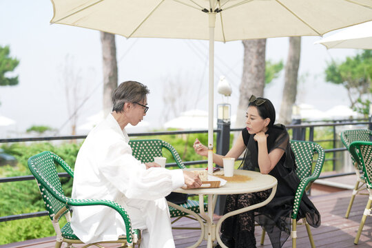 A Korean man and woman in their late 50s enjoy coffee and cake at a seaside cafe terrace with parasols. The scene is calm and peaceful, touched by the early summer breeze. Incheon, South Korea.