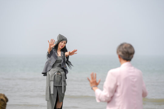 In June 2025, a Korean man and woman in their late fifties are standing and talking beside the rugged shoreline filled with jagged rocks and crashing waves in Incheon Metropolitan City, South Korea.