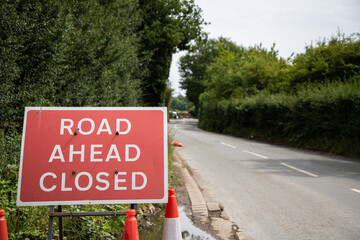 Photo of a red "Road Ahead Closed" sign placed at the roadside. Temporary roadwork or closure warning sign used in the UK, captured in a natural outdoor setting with grass and tarmac visible.