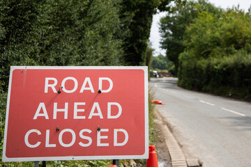 Photo of a red "Road Ahead Closed" sign placed at the roadside. Temporary roadwork or closure warning sign used in the UK, captured in a natural outdoor setting with grass and tarmac visible.