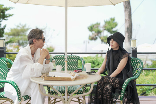 A Korean man and woman in their late 50s enjoy coffee and cake at a seaside cafe terrace with parasols. The scene is calm and peaceful, touched by the early summer breeze. Incheon, South Korea. - Powered by Adobe