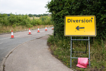 Yellow "Diversion" sign with a black arrow pointing left, positioned by the roadside. UK traffic sign indicating an alternative route due to roadworks or closures. Clear and visible.