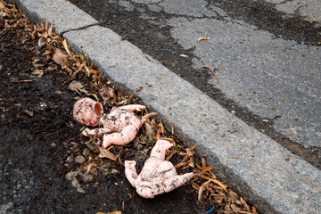 Broken and dirty baby doll lies abandoned on the roadside among dead leaves and mud. A haunting,...