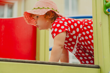 A young child wearing a red polka dot dress and a hat is climbing on colorful playground equipment The sun is shining in a vibrant outdoor park setting creating a joyful atmosphere