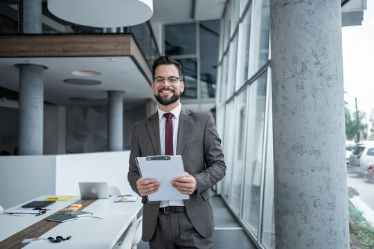 Smiling businessman holding clipboard in modern office - Powered by Adobe