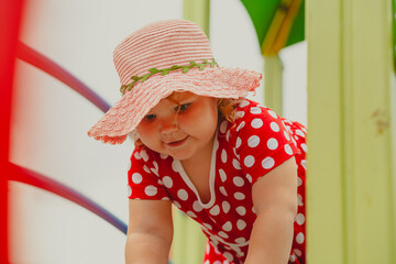 A young child wearing a red polka dot dress and a hat is climbing on colorful playground equipment The sun is shining in a vibrant outdoor park setting creating a joyful atmosphere