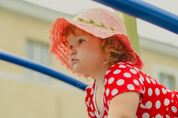 A young child wearing a red polka dot dress and a hat is climbing on colorful playground equipment The sun is shining in a vibrant outdoor park setting creating a joyful atmosphere