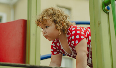 A young child wearing a red polka dot dress and a hat is climbing on colorful playground equipment The sun is shining in a vibrant outdoor park setting creating a joyful atmosphere