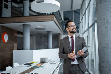 Smiling businessman holding clipboard and pen, looking away in modern office