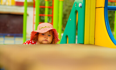 A young child with curly red hair and a pink hat peers out from behind brightly colored playground equipment It is a sunny day perfect for outdoor fun and exploration