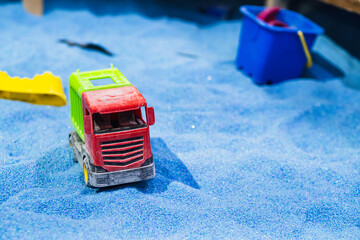 A green and red toy truck drives through vibrant blue sand at a fun childrens play area surrounded by colorful tools and bright sunlight in the afternoon