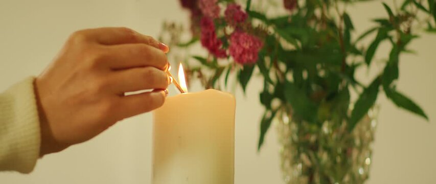 Hand lighting a large white candle with flowers in the background indoors