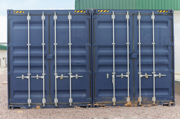 two blue shipping containers parked outdoors in front of a warehouse on the gravel