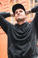 A man stands with hands on his head wearing a black hoodie and cap He looks contemplative in front of a textured red brick wall on a sunny day