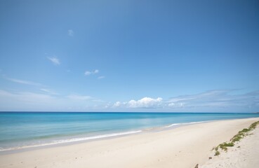 Obraz premium Empty pristine beach with tranquil turquoise ocean waves meeting soft white sand under clear blue sky with scattered clouds. Gentle surf laps shoreline, creating serene coastal landscape perfect for