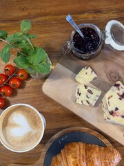 Rustic wooden table displaying fresh pastries, golden brown coffee with intricate latte art, homemade cherry jam, ripe cherry tomatoes, garnished with mint leaves
