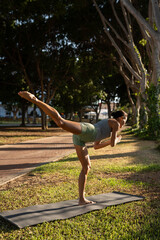 Fit young woman balancing in warrior III yoga pose on a mat in a tranquil park during golden hour,...