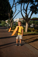 Blond young boy standing on a park path at sunset, holding a bright orange toy airplane in one...