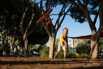 Energetic young blond boy throwing an orange toy airplane in a sunlit park, captured mid-motion...