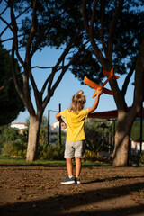Blond young boy in yellow t-shirt and grey shorts standing in a park with back to the camera,...