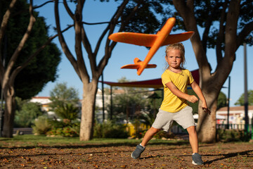 Energetic young blond boy throwing an orange toy airplane in a sunlit park, captured mid-motion...