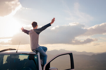 Young man sitting on his car with arms wide open, embracing freedom and nature during a scenic road trip at sunset. © Marcio
