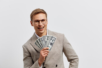 Happy young man wearing glasses holds a fan of hundred-dollar bills and smiles confidently, dressed...