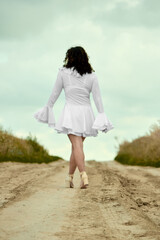 A young woman with curly hair, wearing a white dress and high heels, walks along a dirt road into...