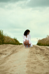 A young woman with dark curly hair, wearing a white dress, sits in a squatting position in the middle of a sandy road, looking thoughtful and contemplative with a serene cloudy sky .