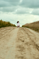 Brunette woman in white shirt crouching on a sandy road amid the fields under a cloudy sky, a moment of serenity and contemplation in the expansive landscape captured in a captivating shot .