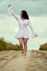 A young woman walks with joy in a white dress and cream shoes down a sandy road with one arm raised up into the air in a joyous manner on a beautiful summer day.