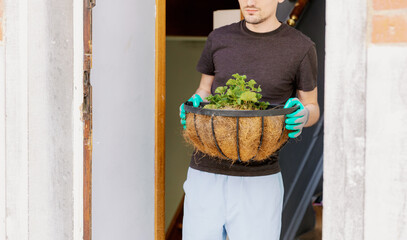 A young man carries a flower in a pot out of the house.
