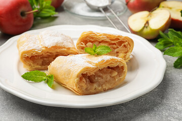 Pieces of tasty apple strudel with powdered sugar, mint and fruits on grey table, closeup