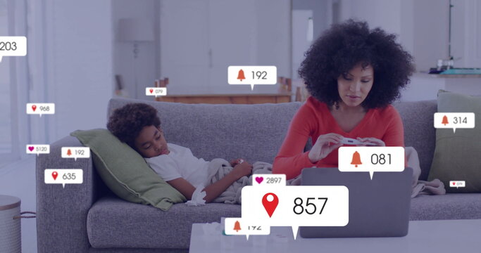 Working mother typing on laptop on sofa in living room, with smartphone, controller and alert icons