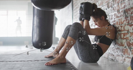 Sitting athlete wearing boxing gloves, leggings by brick wall on gym floor, with heavy punching bag