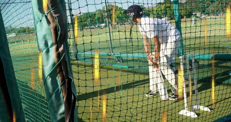 Practicing male cricket batsman in white uniform holding bat at net cage on field, with stumps