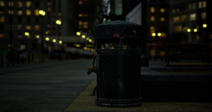 A trash bin sits quietly on a city street, softly illuminated by nearby street lights. The urban landscape shows buildings and emptiness during late evening hours.