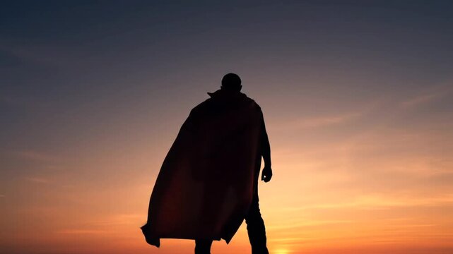 Silhouette of a superman in a cape standing against a vibrant sunset sky at dusk horizon view