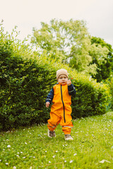 A joyful toddler in a bright orange rain suit explores a lush green garden filled with vibrant...