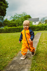 An adorable little toddler, dressed in a bright orange rain suit, joyfully walking outside in a...