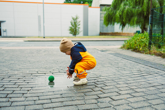 A playful child dressed in vibrant and bright rain gear joyfully exploring the great outdoors with a green ball, happily playing on a wet and shiny cobblestone path after a rainy day