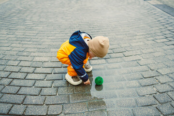A joyful child plays with a bright ball, jumping into rainwater puddles on a charming cobblestone...