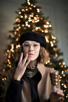 Cute young woman in winter outfit with berett, while wiping mouth after eating holiday treats out of Christmas tree. Funny fashionable woman enjoys eating sweets during X-mas