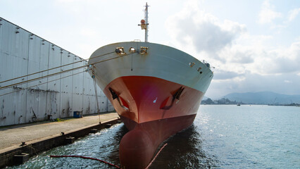 Large cargo ship moored at a port berth, seen from the bow, with industrial buildings and mountains...