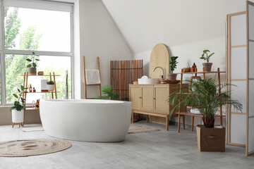Interior of light bathroom with folding screen, bathtub and houseplants