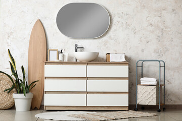 Chest of drawers and wooden surfboard near white grunge wall in interior of bathroom