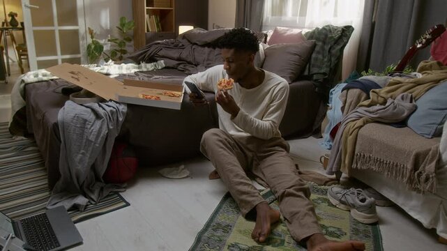 Young African American man sitting on floor, enjoying slice of pizza and looking at smartphone in his messy bedroom with undone bed